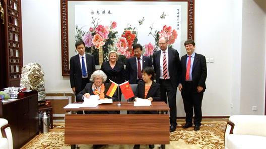 Man sieht einen mit der deutschen und der chinesischen Flagge geschmückten Schreibtisch, an dem zwei Damen sitzen, die das Kooperationsabkoomen unterzeichnen möchten. Hinter ihnen stehen deutsche und chinesische Delegationsmitglieder.